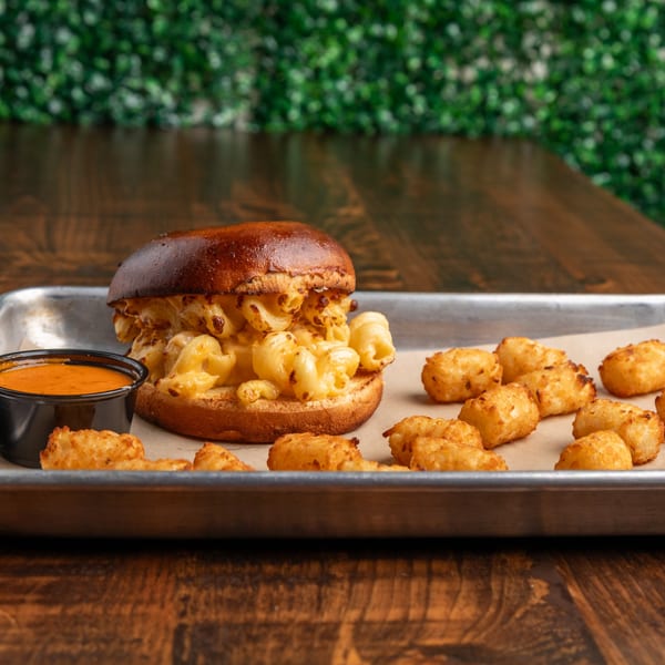 Mac and cheese burger with tater tots and dipping sauce on a metal tray.
